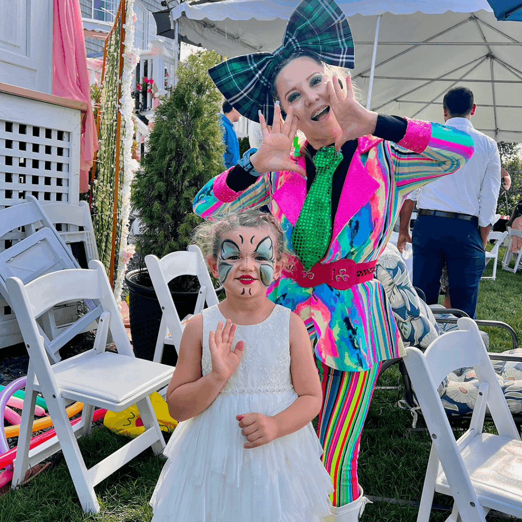 Niña con vestido blanco y maquillaje de mariposa en el rostro, posando junto a una mujer disfrazada con un traje colorido y un gran lazo en la cabeza, en un evento al aire libre.